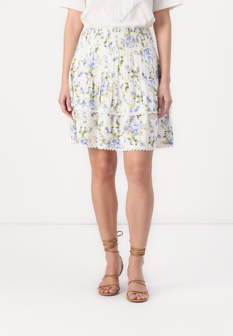 Femme portant une jupe blanche à volants avec des fleurs bleues et vertes et des sandales plates marron à brides, debout sur fond blanc.