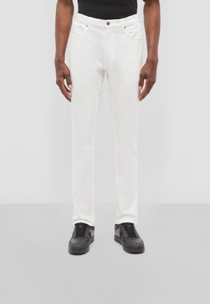 Hombre con pantalones ajustados blancos y zapatillas negras de pie frente a un fondo liso de color gris claro.