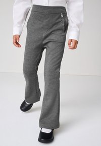 Kind trägt graue Schlaghose, weißes Hemd, weiße Socken und schwarze Schuhe, geht auf einem schlichten weißen Boden und vor weißem Hintergrund.