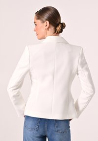 Blazer blanc ajusté avec un col cranté et des épaules structurées, présentant une texture lisse et des détails de couture subtils. Porté avec un jean bleu.