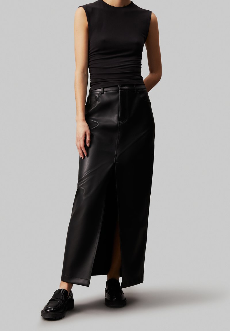 Gonna maxi nera in pelle con spacco frontale, abbinata a un top aderente senza maniche nero e scarpe chunky nere. Texture liscia, design su misura.