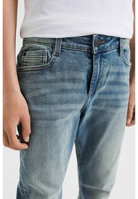 Pantalones vaqueros de denim azul claro con un ligero desgastado, cinco bolsillos y cierre de botón y cremallera. La tela parece suave con una textura sutil.