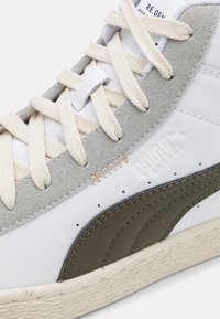 Bílé a šedé tenisky Puma s béžovými tkaničkami, zamilovanou detailní zúženou úpravou, vyraženým logem a texturovanou podrážkou, zobrazené z boční a přední strany.