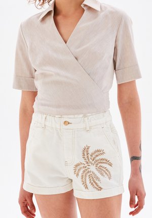 Beige kortärmad omslagblus med en subtil glans, kombinerad med vita shorts med palmbladsbroderi i tan.