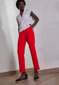Camicia bianca a maniche corte con taschino, abbinata a pantaloni rossi su misura. Scarpe espadrillas nere con suola a zeppa. Texture lisce.