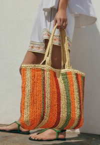 Sac en toile tissé avec des rayures orange, rouge, vert et beige, doté de deux poignées robustes et d'un design texturé et flexible.