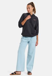 Schwarzer Kapuzenpullover mit Fronttasche und Logo, kombiniert mit hellblauen weiten Jeans und schwarzen Riemchensandalen. Glatte Stofftextur.