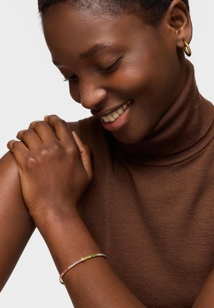 Mujer sonriente con cuello alto marrón sin mangas, lleva un pendiente de aro dorado y una pulsera con gemas multicolores, mirando hacia abajo con las manos suavemente cruzadas.