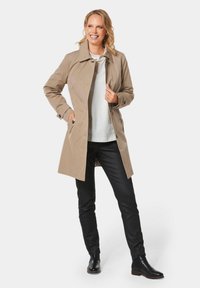 Beige trenchcoat met een kraag, knoopdetails en zijzakken, gecombineerd met een witte blouse en zwarte broek, compleet met zwarte enkellaarsjes.