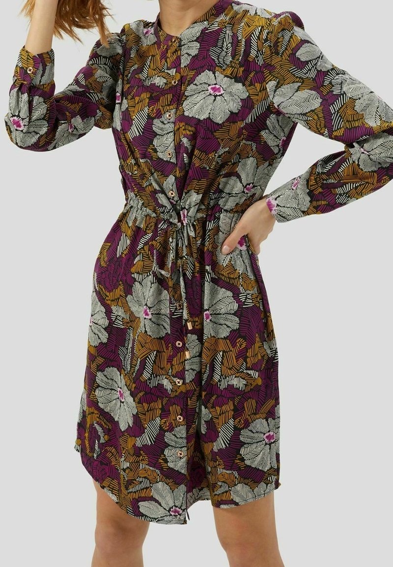 Femme portant une robe chemise à manches longues avec un devant à boutons et un motif floral en violet, moutarde, blanc et noir, cintrée à la taille.