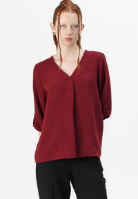 Burgunderfarbene Bluse mit V-Ausschnitt, strukturiertem Stoff und drei Viertel langen hochgerollten Ärmeln. Besitzt eine Frontfalte und Knopfdetails an den Ärmeln.