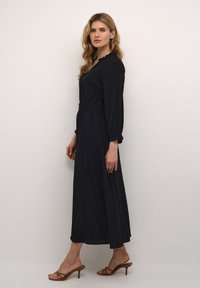 Ilga juoda suknelė su ilgomis rankovėmis, apkabinta talija, V formos iškirpte ir subtiliu tinklelio raštu; dėvima su aukštakulniais sandalais. Lygus audinio paviršius.