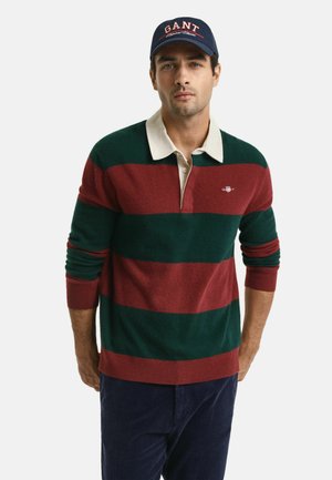 Grüner und burgunder gestreifter Wollpullover mit einem beigen Kragen, ausgestattet mit einem kleinen Logo auf der Brust, kombiniert mit dunklen Cordhosen und einer marineblauen Mütze.