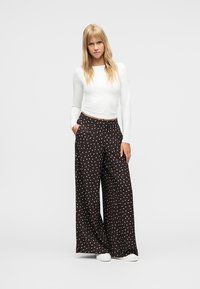 Νεαρή γυναίκα με μακριά ξανθά μαλλιά, που φοράει λευκό μακρυμάνικο τοπ, μαύρο παντελόνι φαρδύ με λευκά πουά και λευκά παπούτσια, όρθια.