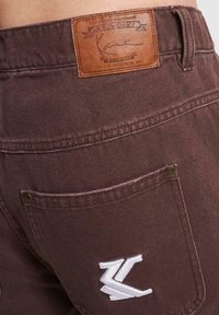Brune denimshorts med et lærpatch med embossert etikett og en hvit brodert logo på baklommen.