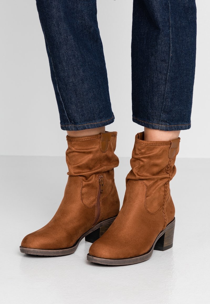 Stivaletti alla caviglia in suede marrone con un design morbido, cerniera laterale e tacco impilato. Presentano dettagli intrecciati e una texture liscia.