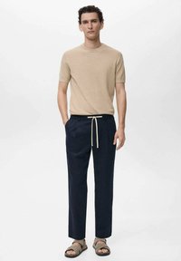 Beige korte mouw gebreid shirt gecombineerd met donkerblauwe taps toelopende broek met een drawstring taille en bruine sandalen, weergegeven tegen een neutrale achtergrond.