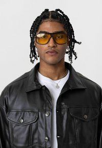 Zwarte leren jas met drukknoopsluiting, gedragen over een wit shirt. De persoon draagt geel getinte zonnebrillen en meerdere zilveren kettingen.