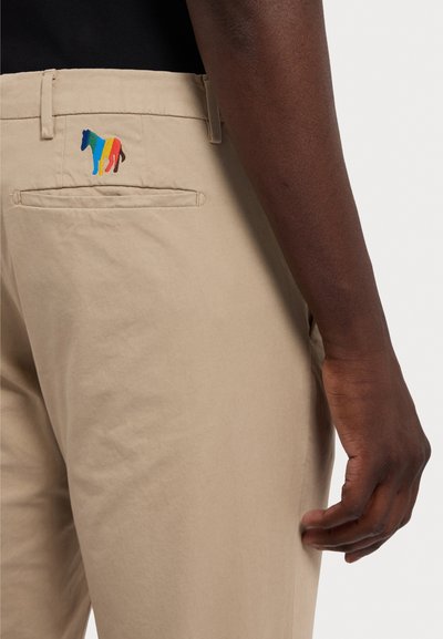 Chinos bege com um corte slim, apresentando um bordado colorido de um cavalo no bolso de trás e uma textura de algodão suave.