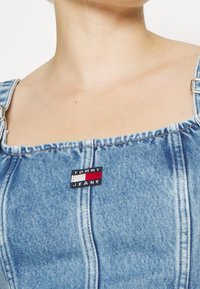 Denimowa bluzka z regulowanymi ramiączkami w jasnoniebieskim kolorze, z kwadratowym dekoltem i naszywką z logo "TOMMY JEANS" w kolorze czarnym, czerwonym i białym.