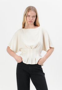 Blusa corta color crema a maniche corte con scollatura ampia e dettaglio in vita plissettato, abbinata a pantaloni neri a vita alta. Tessuto morbido.