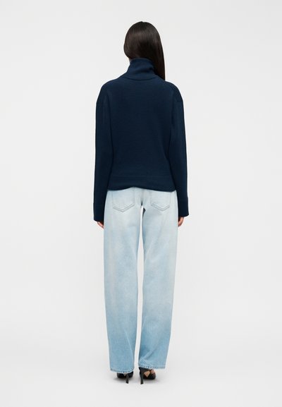 Pull à col roulé bleu marine à manches longues, associé à un jean à taille haute bleu clair à jambes larges. Silhouette ajustée et texture douce.