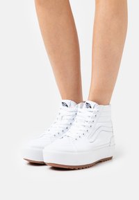 Zapatillas altas blancas de lona con suela de goma, que presentan un diseño de textura en la suela, cordones blancos y un logotipo azul en contraste en la lengüeta.
