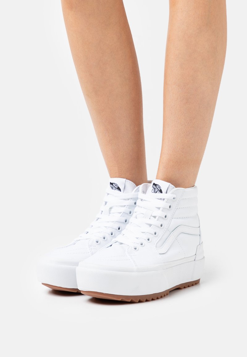 Zapatillas altas blancas de lona con suela de goma, que presentan un diseño de textura en la suela, cordones blancos y un logotipo azul en contraste en la lengüeta.
