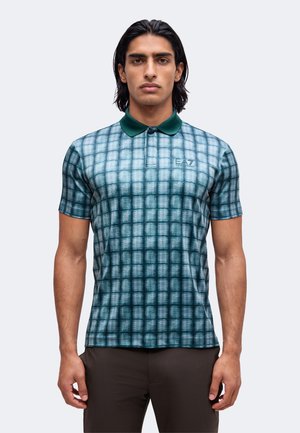 Mann mit langen dunklen Haaren trägt ein blau-grün kariertes Poloshirt und dunkle Hose, steht vor einem einfarbigen hellen Hintergrund.