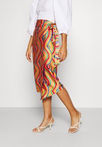 Jupe enveloppante colorée avec un motif ondulé en rouge, bleu et jaune. Fabriquée en tissu lisse, elle présente une taille nouée et une longueur midi.