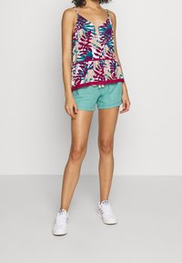 Květinový peplum top v růžové, tyrkysové a bílé barvě, doplněný světlými tyrkysovými šortkami. Modelka má na sobě bílé tenisky.