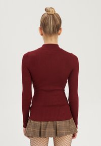 Hranatý top s dlouhými rukávy v burgundské barvě, s nepravým límcem a přiléhavým střihem, z pohledu ze zad, v kombinaci s hnědou kostkovanou skládací sukní.