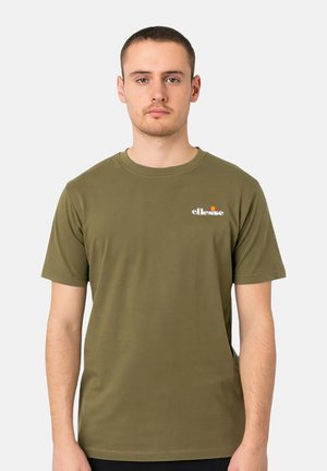 Jeune homme aux cheveux courts portant un t-shirt ellesse vert olive, debout devant un fond uni gris clair.