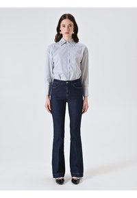 Donna in piedi che indossa una camicia a righe con bottoni infilata in jeans svasati blu scuro, con scarpe nere verniciate su uno sfondo neutro.