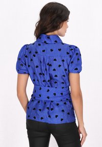 Blusa blu con maniche a sbuffo, decorata con motivi a cuore neri, con vita annodata e tessuto texturizzato. La vista posteriore mostra un dettaglio arricciato sulle spalle.