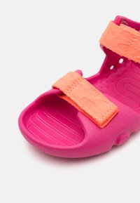 Pink children's sandal made of flexible material, featuring an orange strap with a textured surface and a contoured sole with ridges.