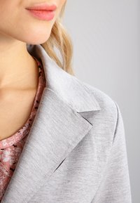 Blazer gris clair au toucher doux, doté d'un col cranté et de coutures détaillées, superposé à un blouse à motifs floraux.