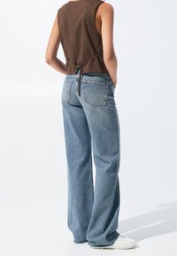 Personne portant un jean bleu clair ample, des baskets blanches et un haut marron sans manches avec un détail noué à l'arrière, debout devant un fond blanc.