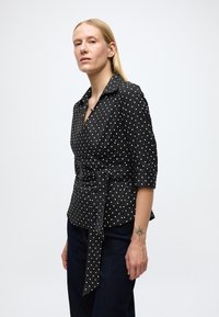 Zwarte wikkelblouse met witte stippen, een kraag, driekwart mouwen en een strikceintuur. De stof oogt glad en heeft een subtiele glans.