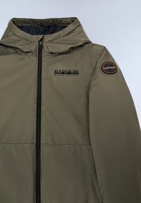 Olivgrön jacka med dragkedjestängning, huva och en strukturerad väv. Den har en svart logotyp på bröstet och en rund applikation på ärmen.