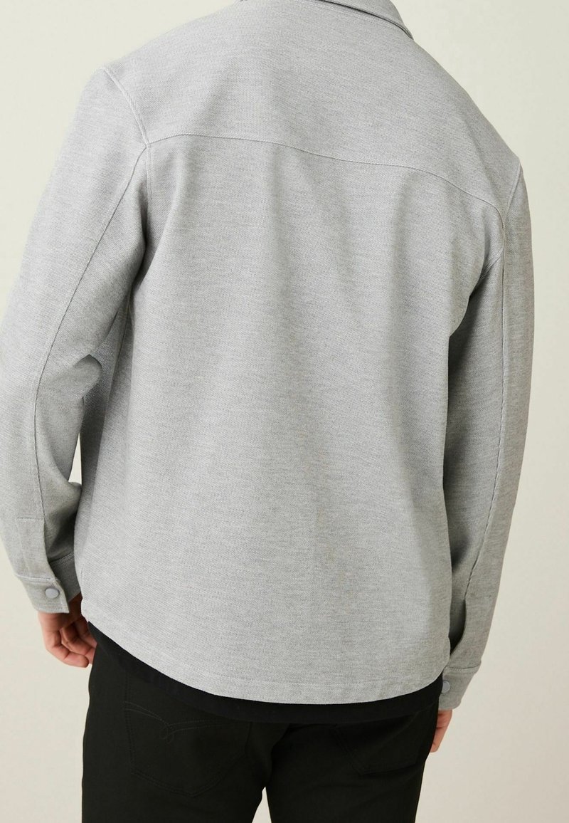 Personne portant une chemise à manches longues gris clair avec des poignets boutonnés, vue de dos sur un fond clair uni.