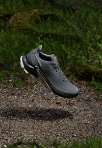 Chaussure de sport gris avec un upper texturé, un design à lacets et une semelle noire contrastante dotée d'accents rouges, positionnée au-dessus d'une surface de gravier.