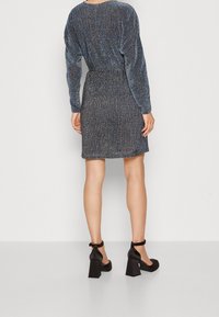 Robe à manches longues bleu scintillant avec taille élastique, ourlet plus court à l'arrière, associée à des chaussures à talons carrés noires avec brides à la cheville.