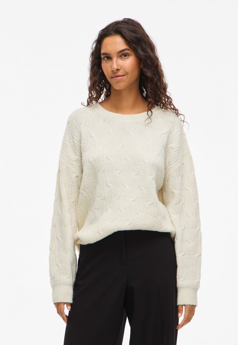 Γυναίκα με σγουρά σκούρα μαλλιά, ντυμένη με ένα κρεμ ζακάρ πουλόβερ και μαύρο παντελόνι, στάθηκε μπροστά σε λευκό φόντο.