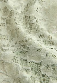 Tissu délicat en dentelle florale blanc cassé avec un maillage complexe et des motifs floraux en relief, légèrement drapé pour montrer la texture et les détails.