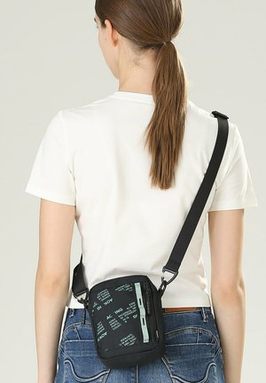 Vrouw met bruin haar in een paardenstaart, gekleed in een wit T-shirt en blauwe spijkerbroek, met een zwarte crossbodytas met groene tekst en rits.