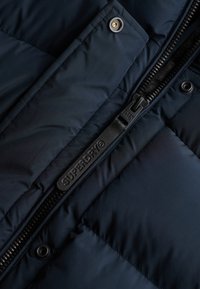 Sezione di giacca imbottita blu navy con texture trapuntata, presenta una cerniera nera con tirante marchiato e dettaglio di bottone a pressione dorato.