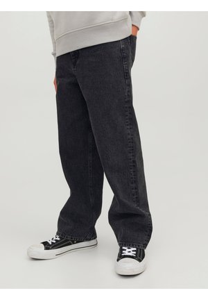 Personne portant un jean gris foncé baggy et des baskets noires et blanches, debout avec une main dans la poche sur un fond uni.