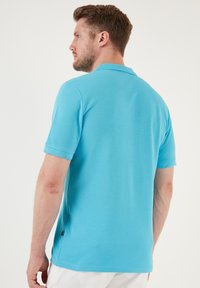 Camisa polo turquesa de algodón. Manga corta, collar de canalé, aberturas laterales y una etiqueta en el dobladillo. Se muestra vista trasera plana.