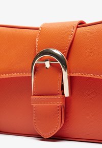 Gros plan d'une sangle de sac en cuir texturé orange avec boucle en argent et coutures visibles sur fond blanc.
